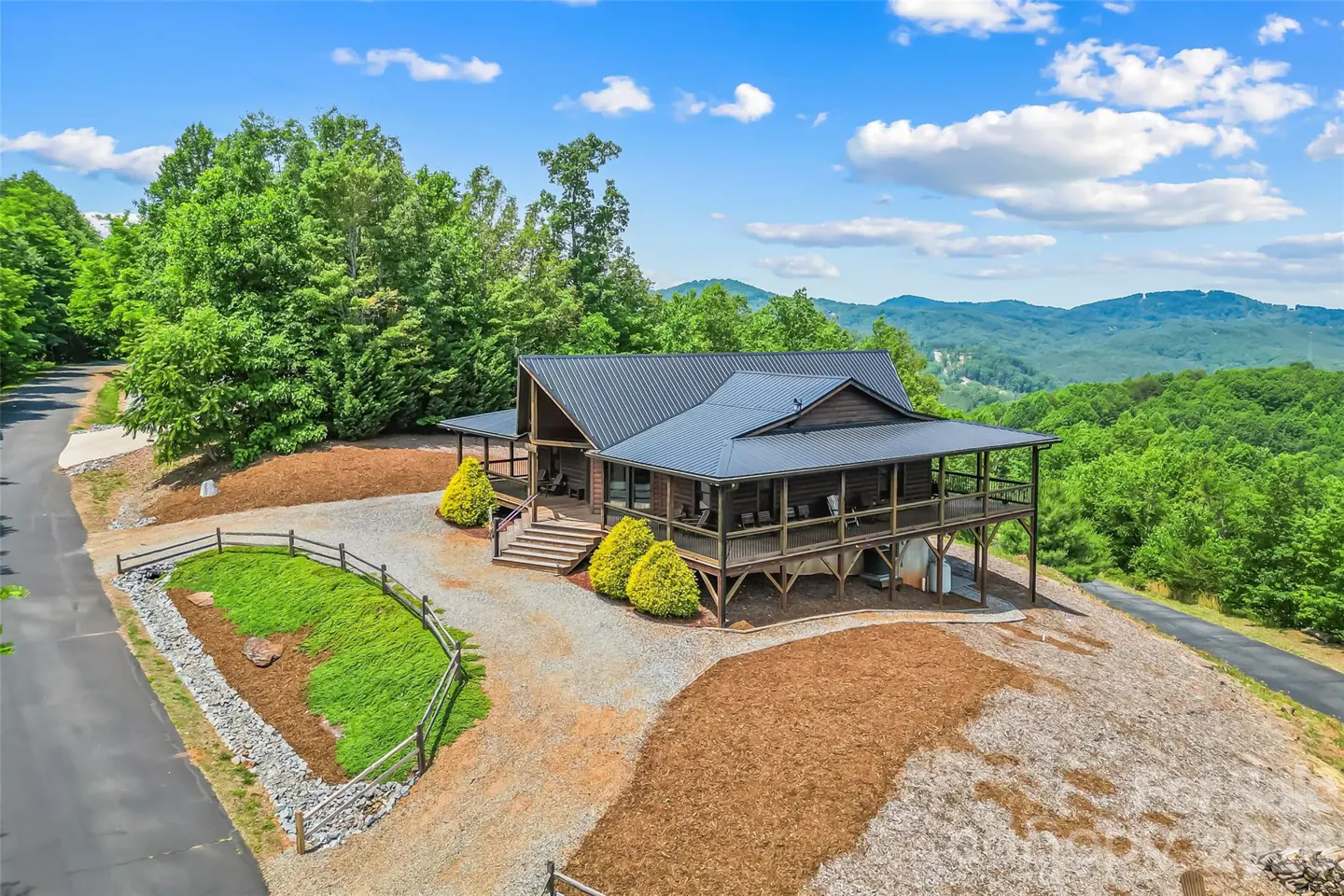 Exterior view of a two-story log cabin with a metal roof, surrounded by trees and mountains. The cabin has a large porch and a gravel driveway.