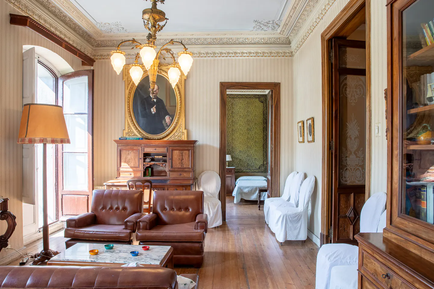 A vintage living room with brown leather chairs, a wooden desk, and a portrait above it. A chandelier hangs from the ornate ceiling.