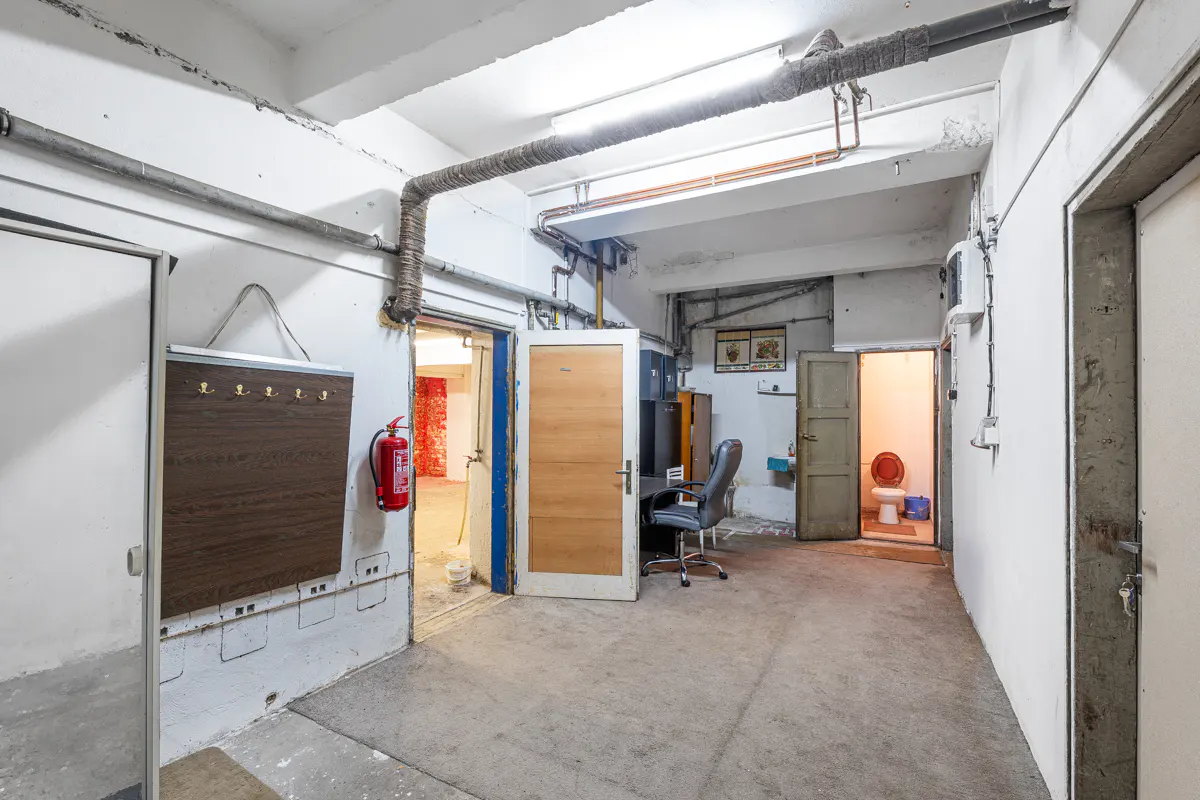 A cluttered basement with white walls, pipes on the ceiling, and multiple open doorways. A desk, chair, and fire extinguisher are visible.