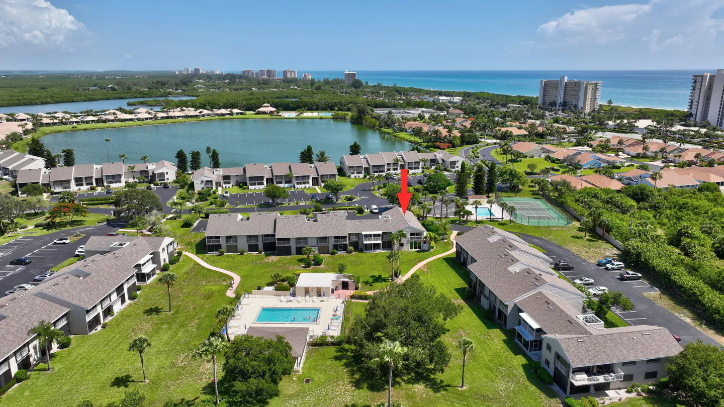Aerial view of a condo complex with a pool, lake, and ocean in the background. An arrow points to a specific unit.