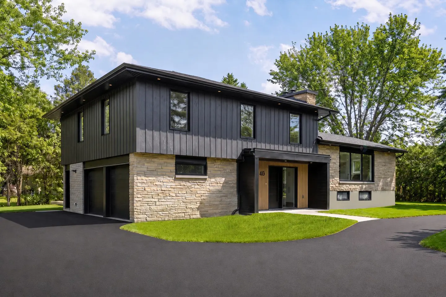Two-story home with black siding, stone facade, and black asphalt driveway. Green lawn and trees surround the house.