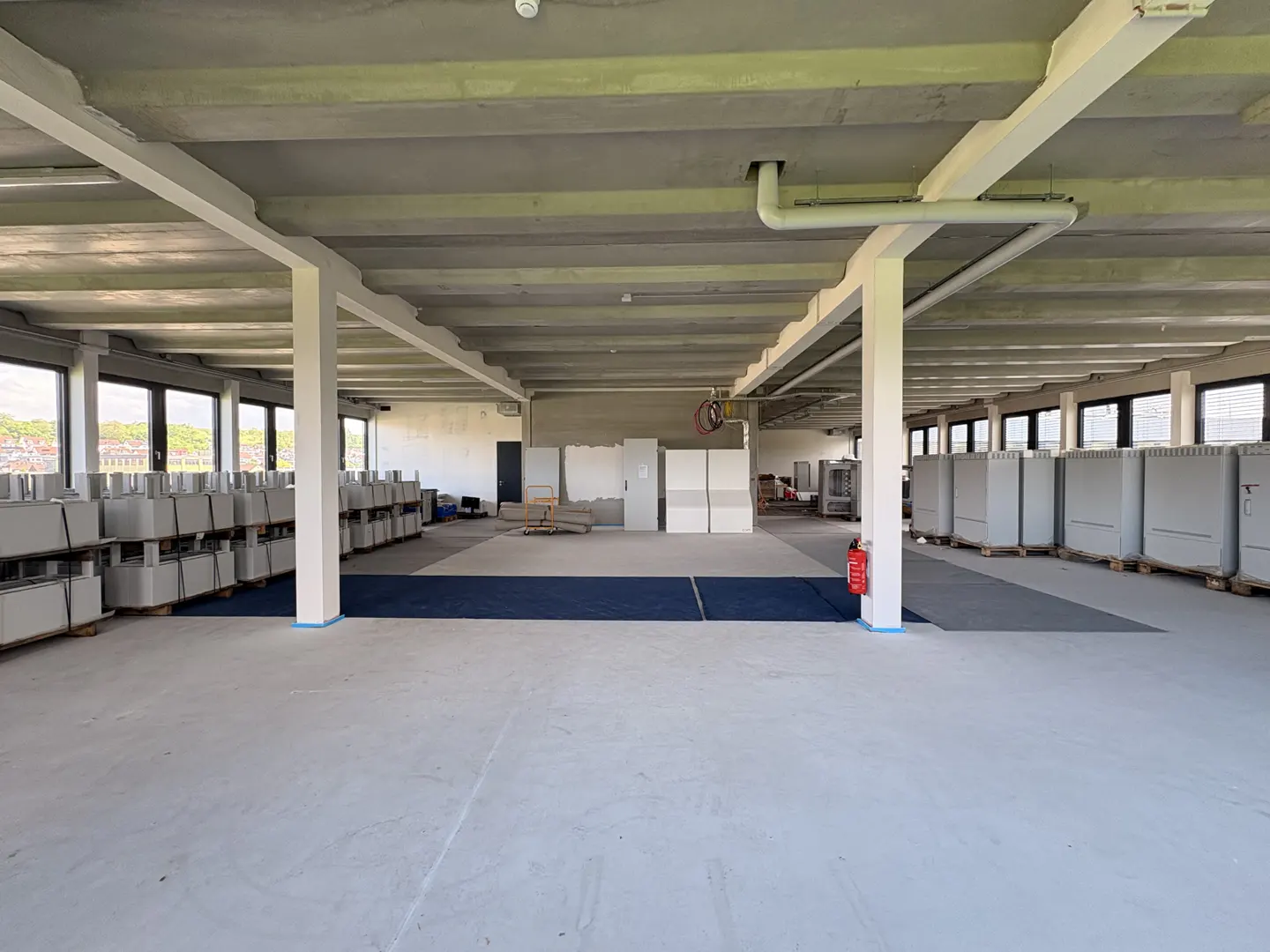 A large, empty commercial space with concrete floors, white pillars, and rows of gray units on pallets. Windows line the left wall, and a red fire extinguisher is visible.
