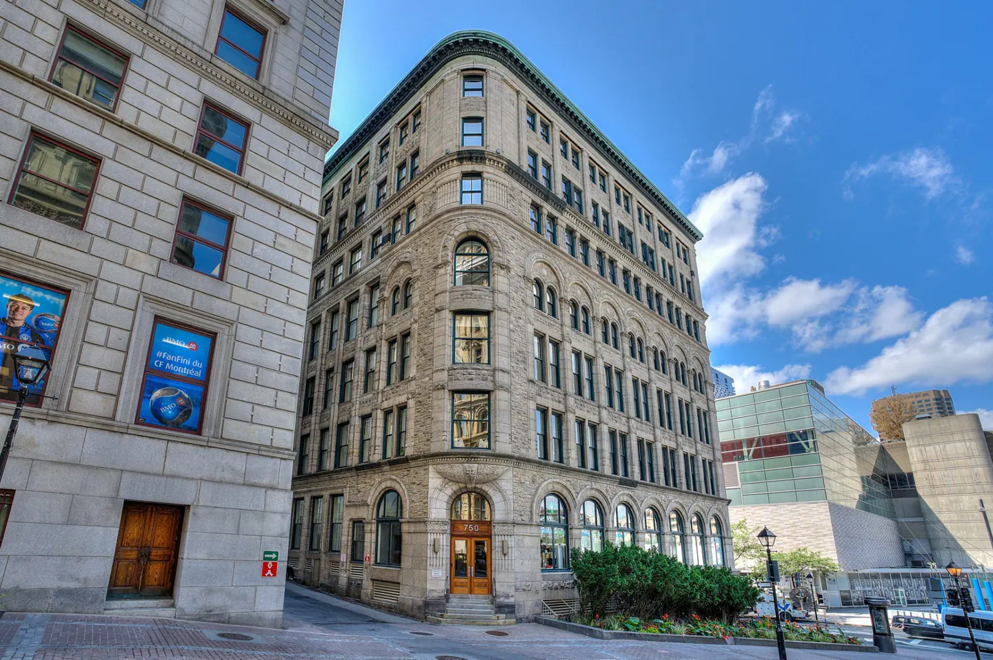 Exterior view of a tall, light-stone building with arched windows, next to a building with sports posters, under a blue sky.