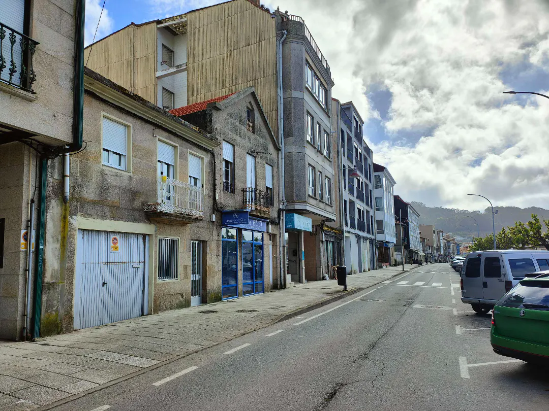 Street view of mixed-use buildings in a European town under a cloudy sky. Cars are parked along the right side of the street.