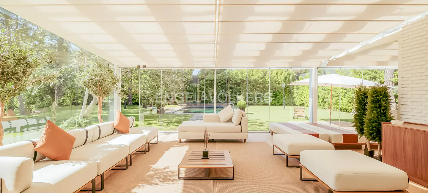 Bright sunroom with white sofas, orange pillows, and a wood coffee table. Glass walls overlook a green lawn, pool, and trees.