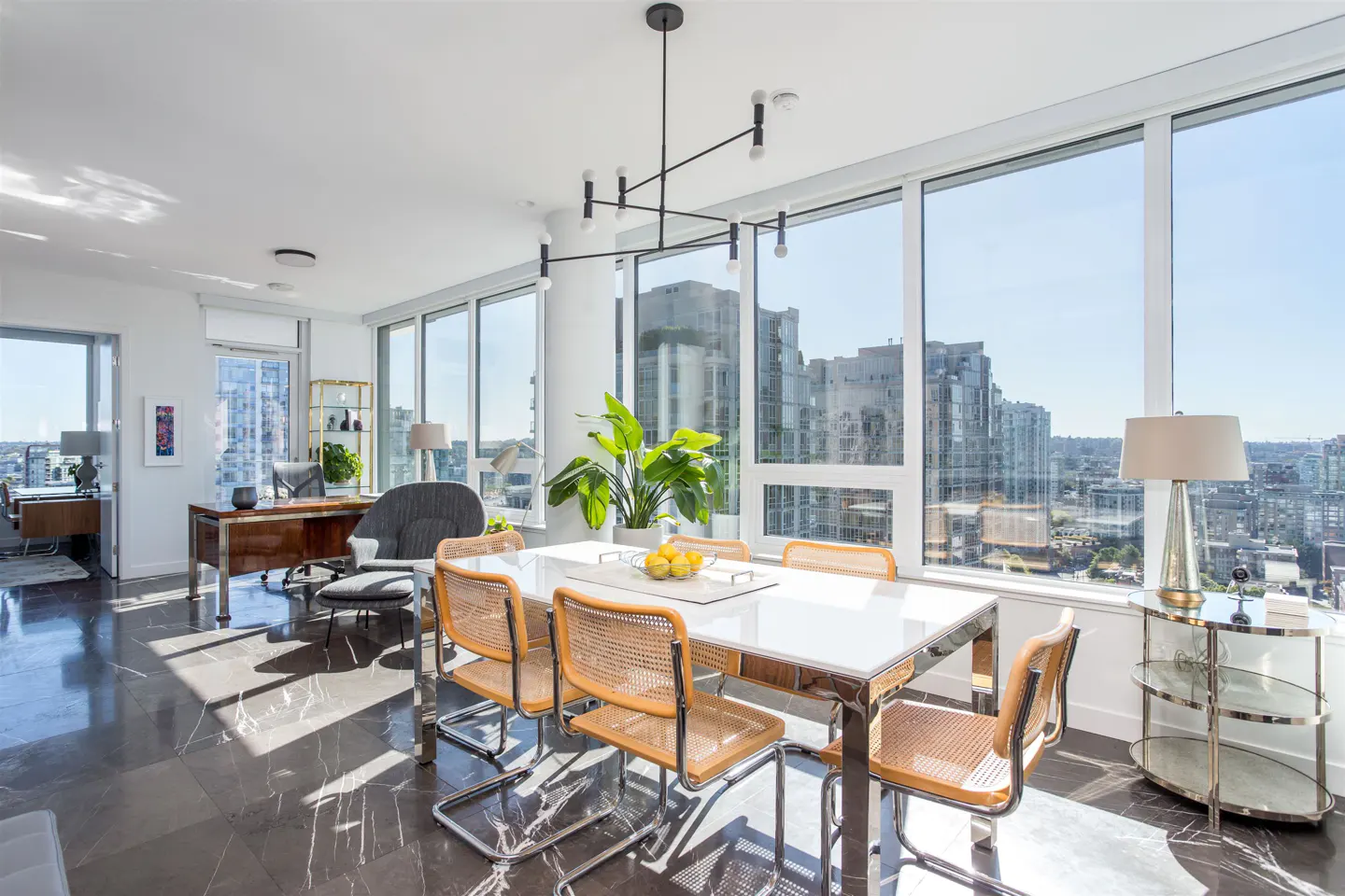 Bright, modern dining area with a white table, wicker chairs, and a black chandelier. Large windows offer a city view.
