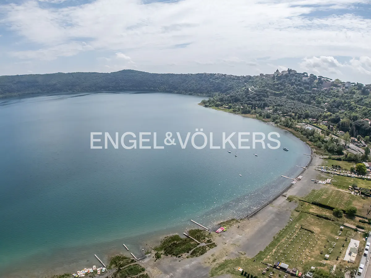 Aerial view of Lake Albano, Italy, with green hills surrounding the blue water. A beach and town are visible on the right side.