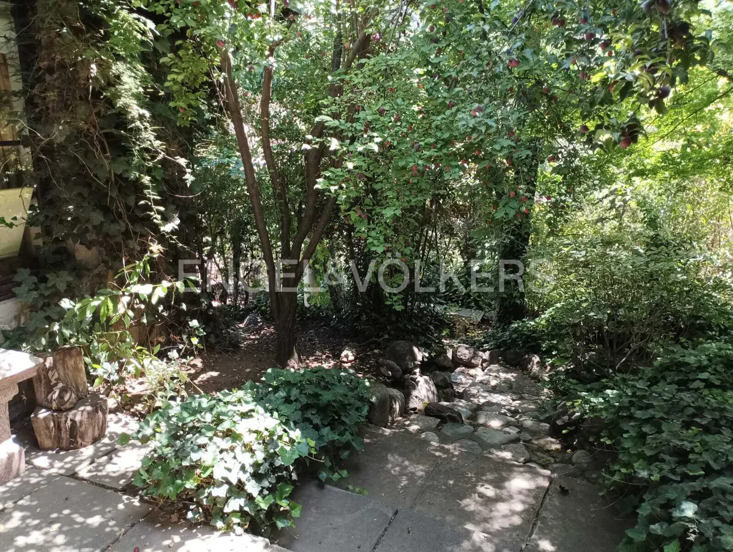 Lush garden with stone path, green foliage, and trees with red fruit. Engel & Völkers logo overlay.