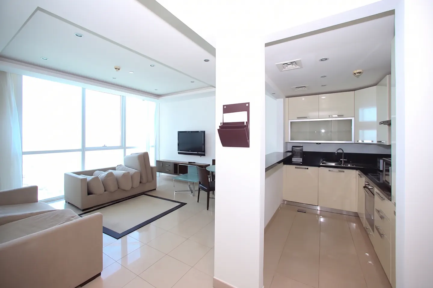 Bright apartment interior with a living room featuring a beige sofa, TV, and a kitchen with cream cabinets and black countertops.
