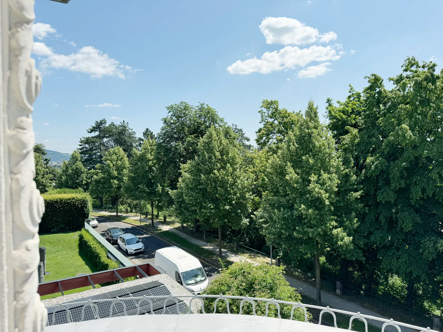View from a balcony overlooking a street with cars, a white van, and lush green trees under a blue sky with scattered clouds.