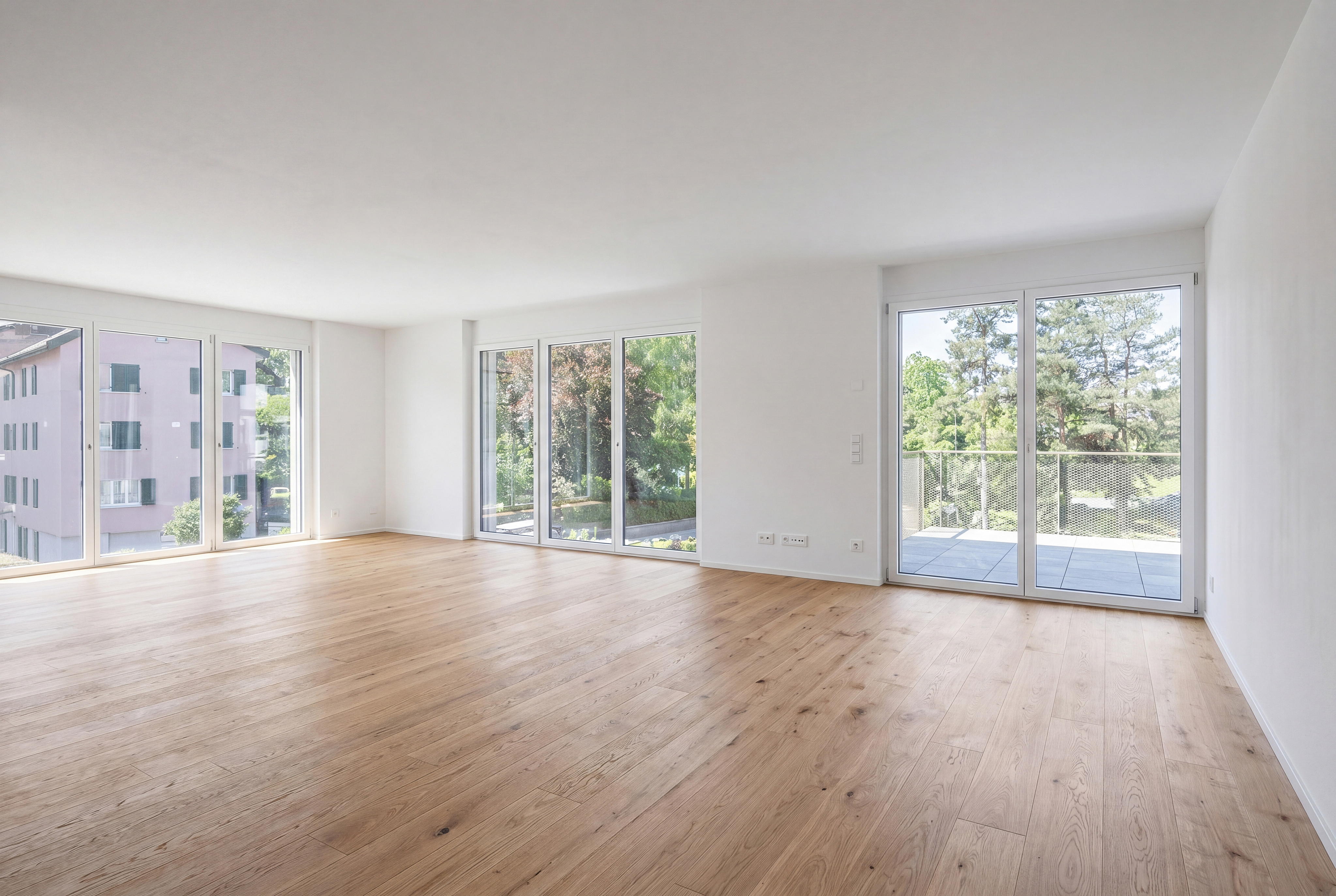 Bright, empty room with light wood floors and white walls. Large windows offer views of trees and a building. [AI-generated text]