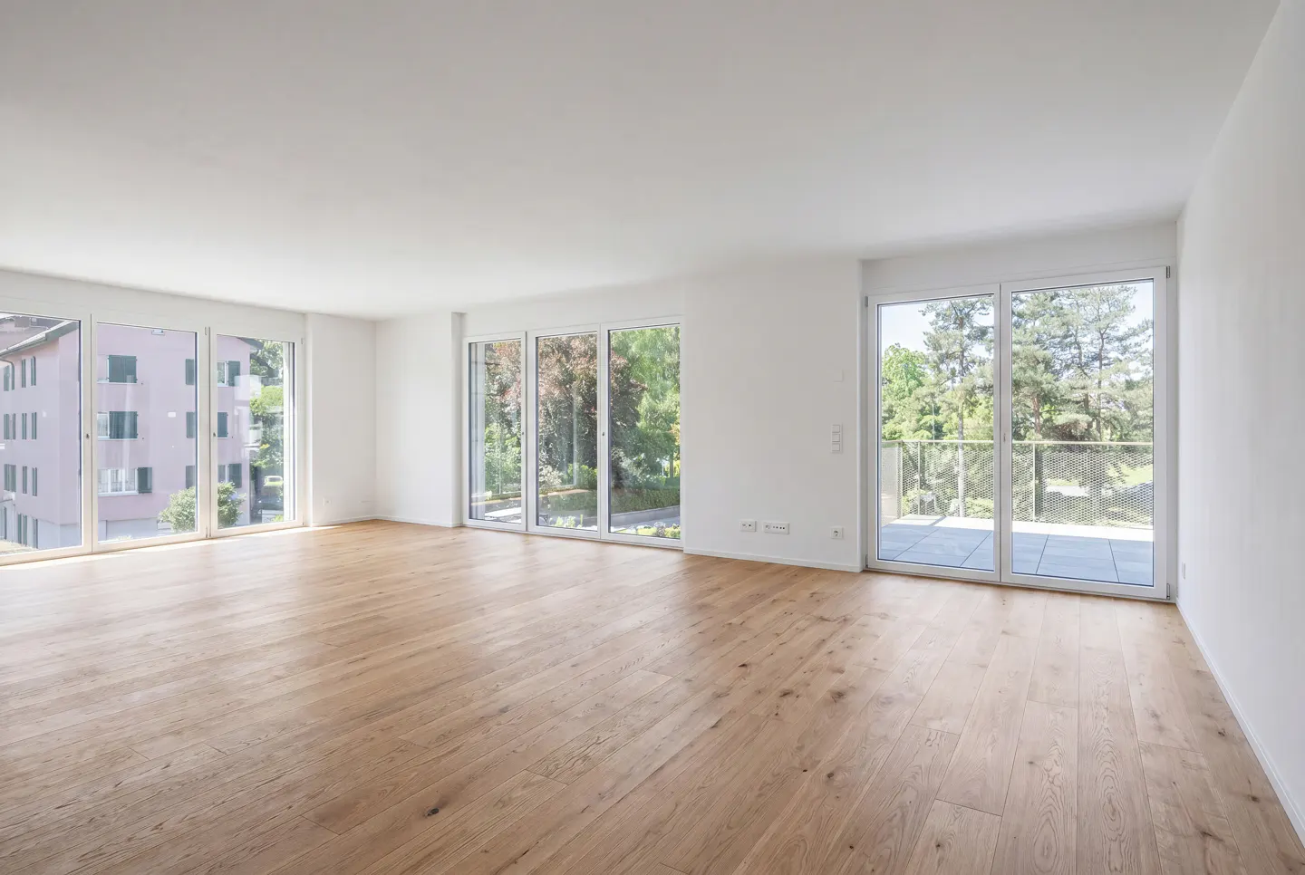 Bright, empty room with light wood floors and white walls. Large windows offer views of trees and a building.