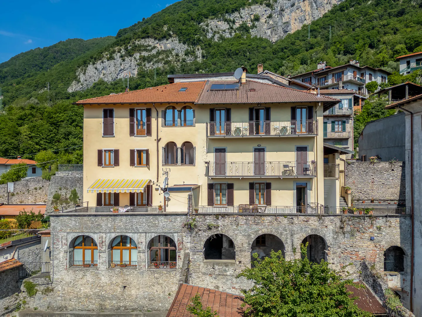 A yellow multi-story building with balconies and a stone arched base, set against a lush green mountain backdrop.