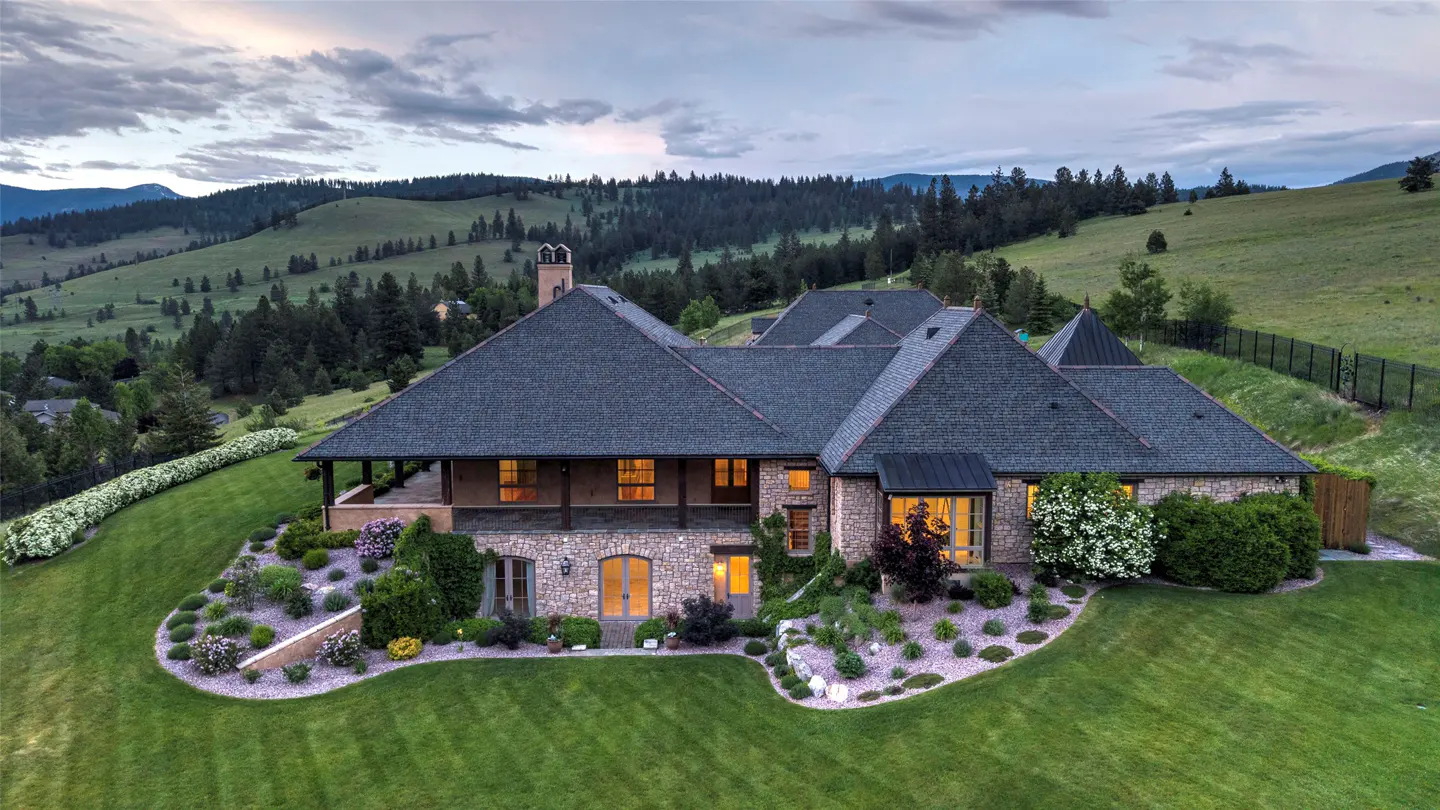 Aerial view of a stone house with a dark roof, surrounded by lush green lawns and landscaping, set against mountain hills.