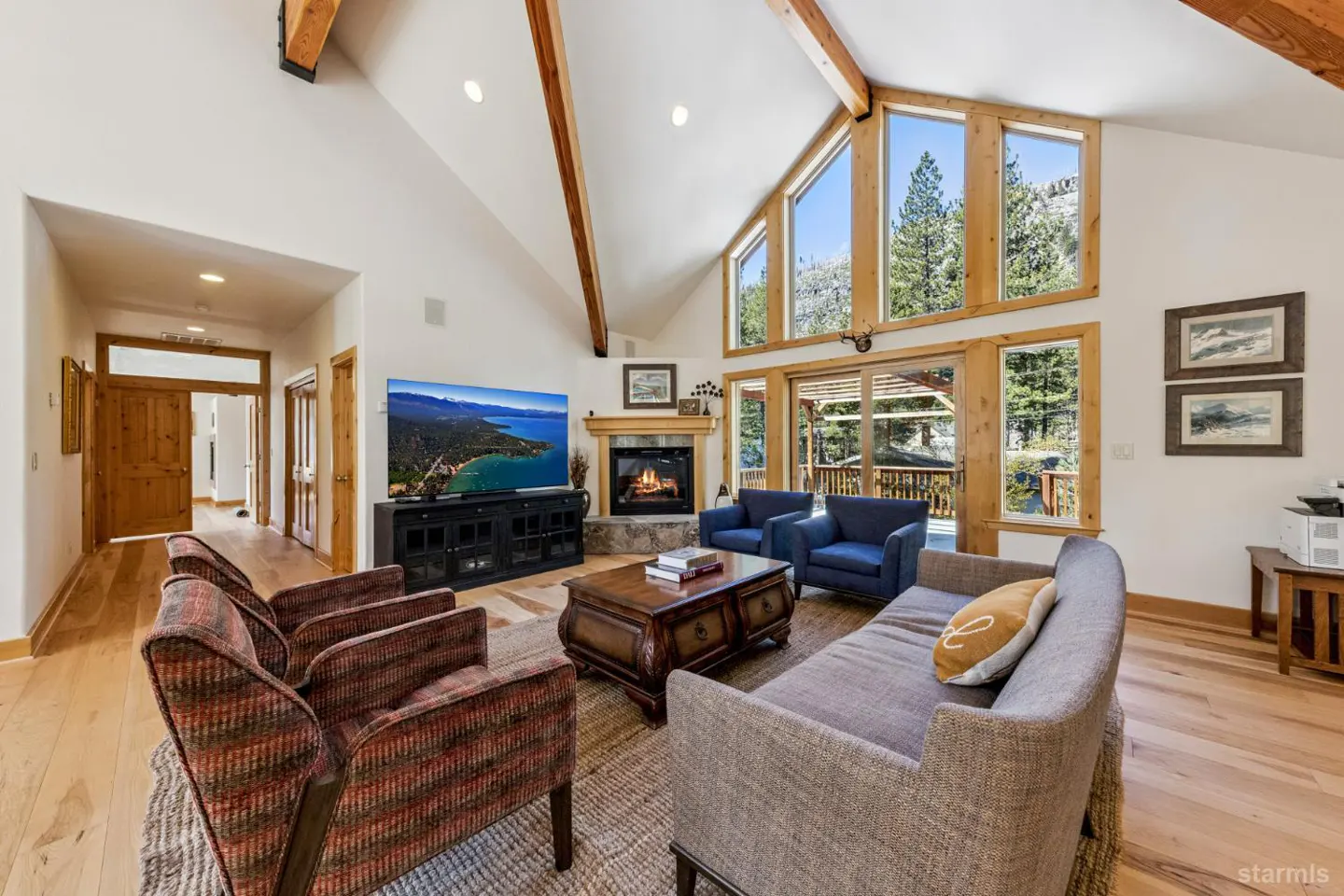 Living room with vaulted ceiling, wood beams, and large windows overlooking trees. A fireplace, TV, sofas, and chairs furnish the space.