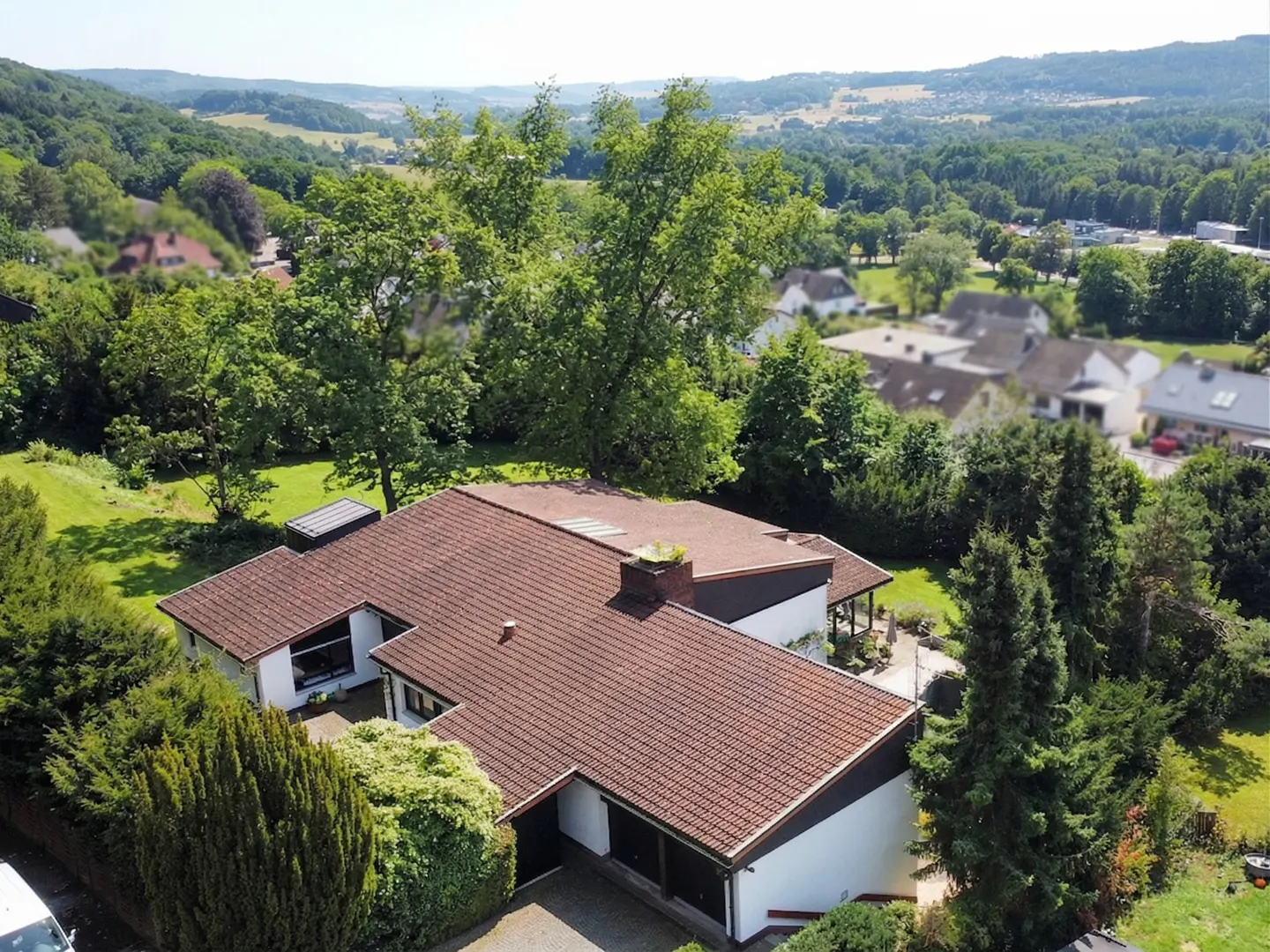 Aerial view of a modern white house with a brown tile roof, surrounded by lush green trees and a distant view of rolling hills.