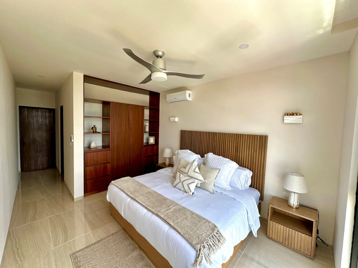 A modern bedroom with a king bed, wood headboard, white linens, and a beige throw. A built-in wood closet and a ceiling fan are also visible.