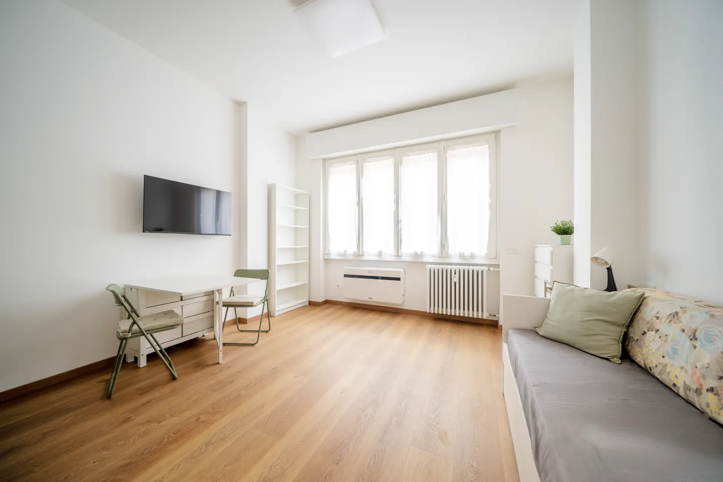 Bright, modern studio apartment with wood floors, white walls, and large window. A TV hangs above a small table and chairs. A daybed sits to the right.