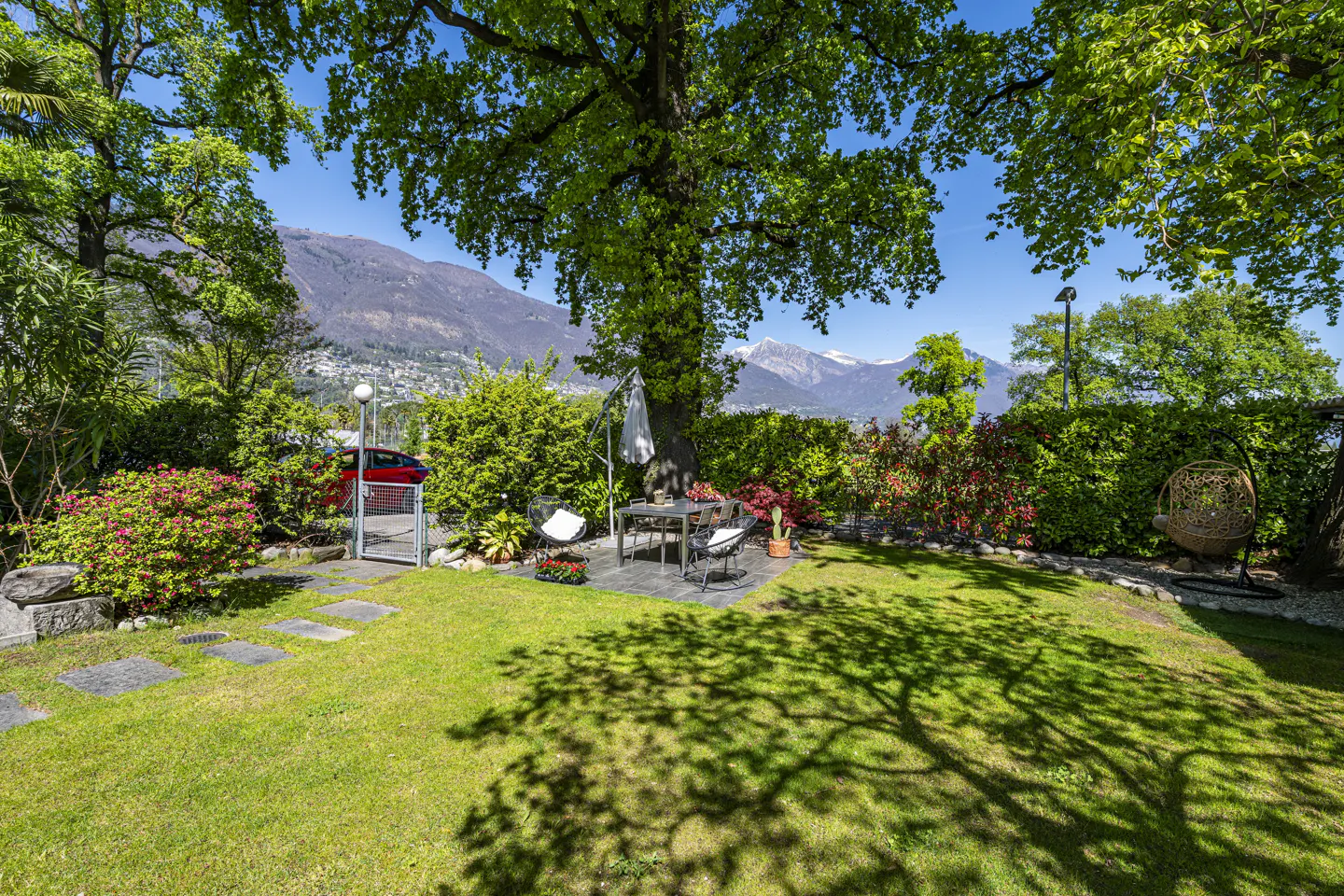 Lush green lawn with a dining set under a large tree, mountains in the background, and a red car parked near a gate.