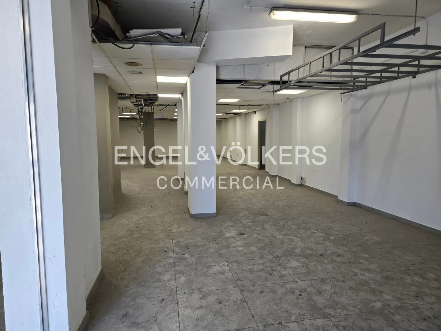 Empty commercial space with white walls, gray floors, and support columns. Fluorescent lights and metal framework hang from the ceiling.