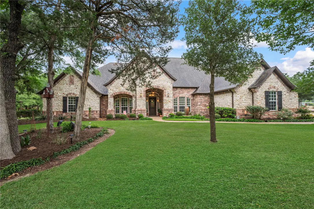 A single-story stone and brick house with a large green lawn and trees. The house has a gray roof and black shutters.
