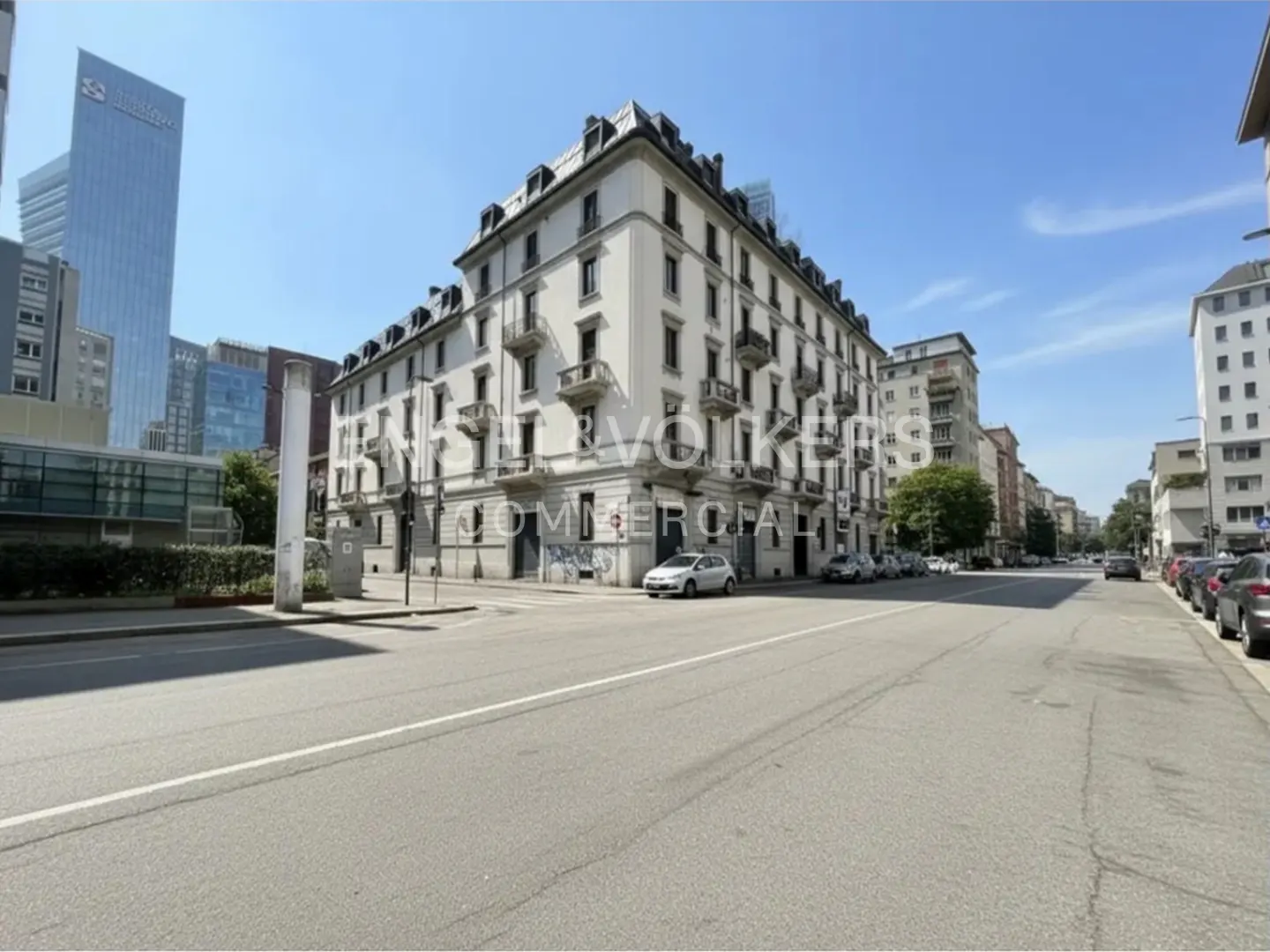 Street view of a white, multi-story building with balconies, cars parked along the side, and a tall skyscraper in the background.