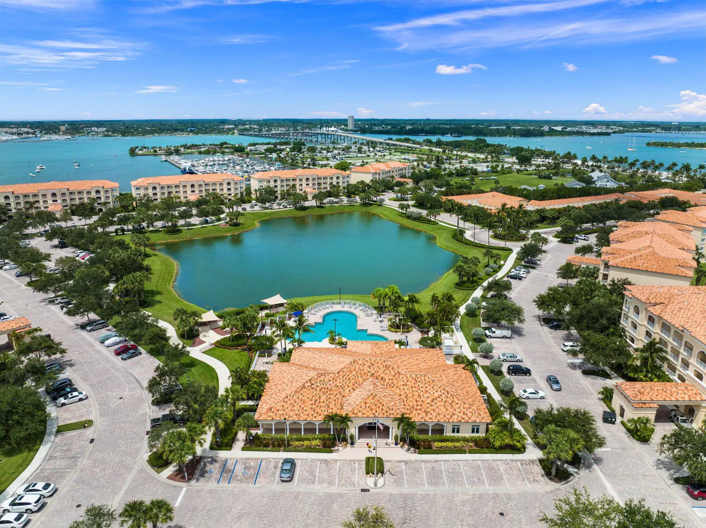 Aerial view of a resort community with a lake, pool, buildings with orange tile roofs, and a bridge in the distance.