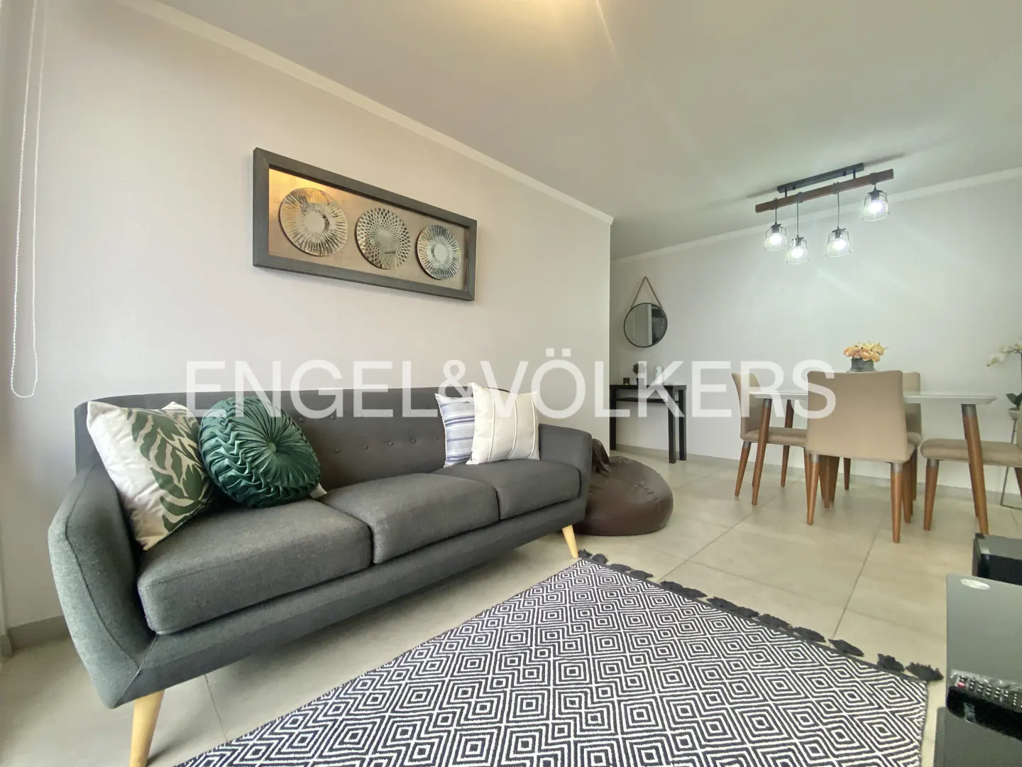 Living room with gray sofa, patterned rug, and dining area. Artwork hangs above the sofa. Neutral color scheme.