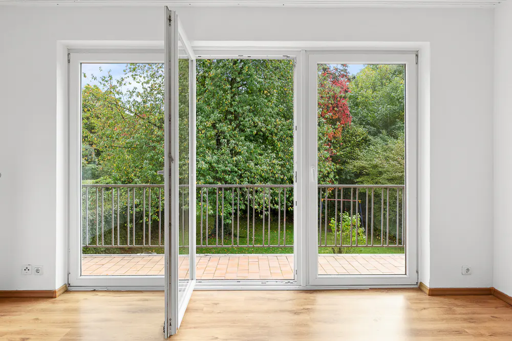 Bright room with wood floor and white walls. Open white glass doors lead to a balcony with green trees.