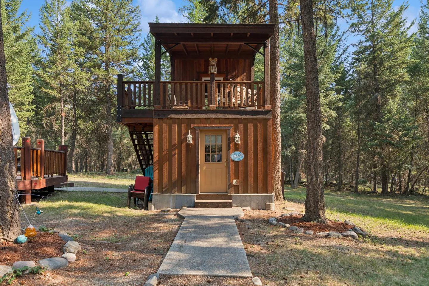 Two-story brown cabin with a balcony and a covered porch, surrounded by tall green trees. A concrete path leads to the front door.
