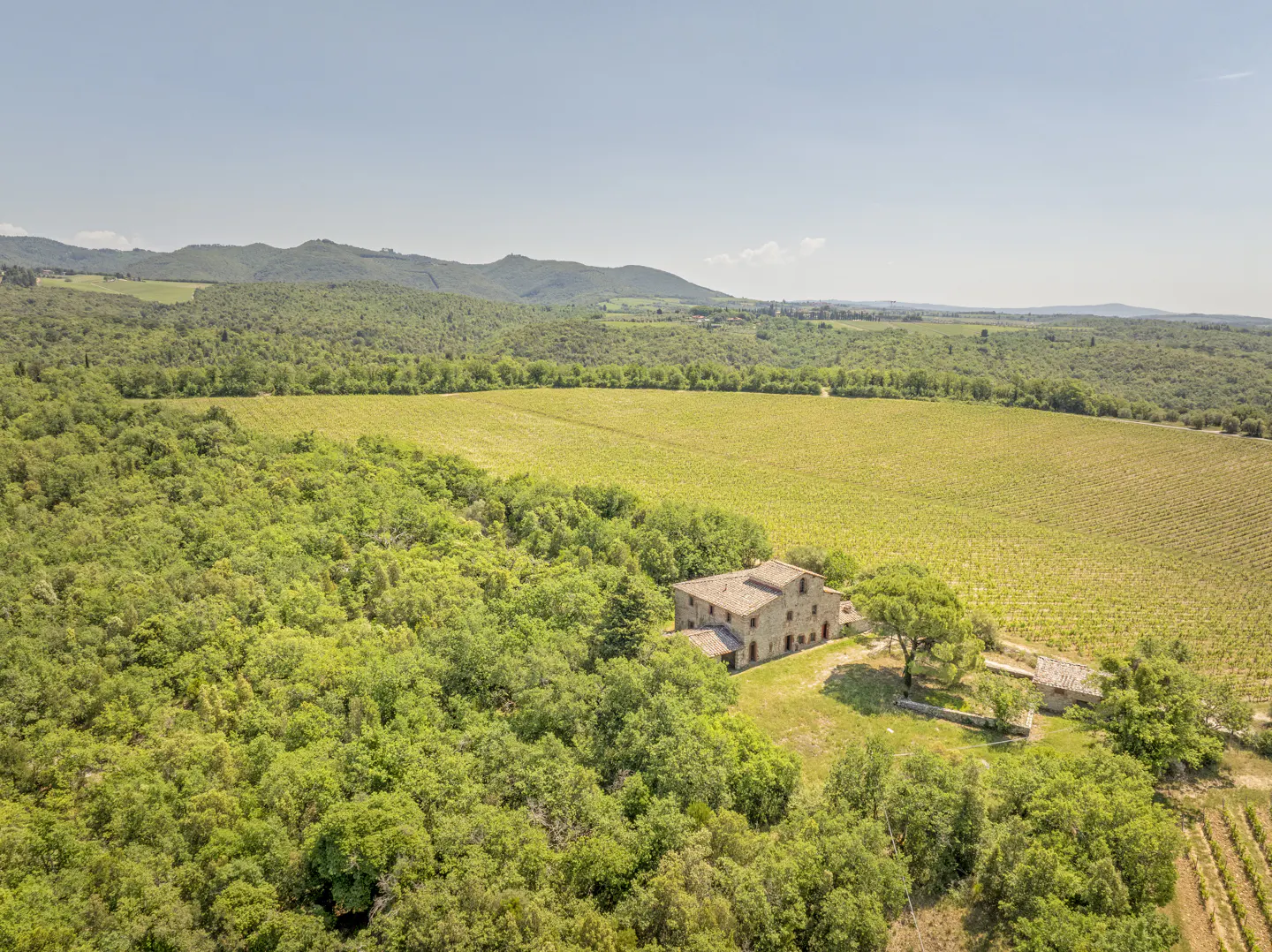 Aerial view of a stone farmhouse nestled in a lush green forest next to a large vineyard with rolling hills in the distance.