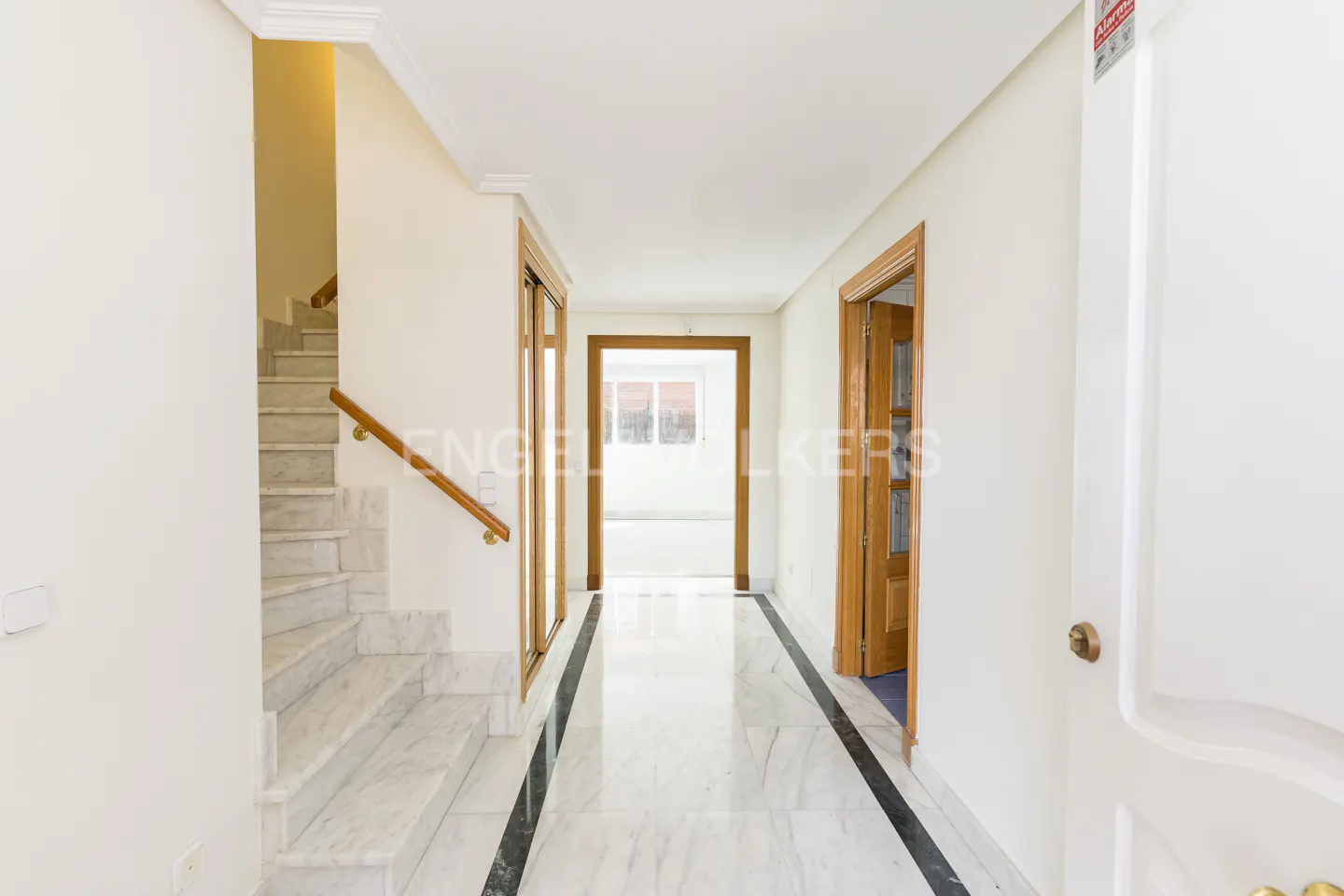 Bright hallway with marble floors, white walls, and wooden doors. A marble staircase with a wooden handrail is on the left.