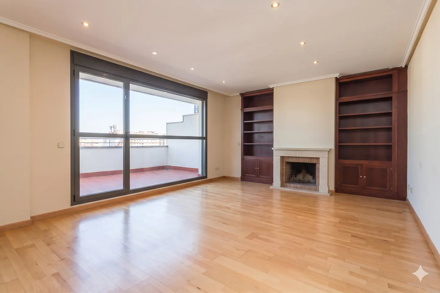 Bright, empty living room with wood floors, fireplace, and built-in dark wood bookshelves. Large sliding glass doors lead to a balcony.