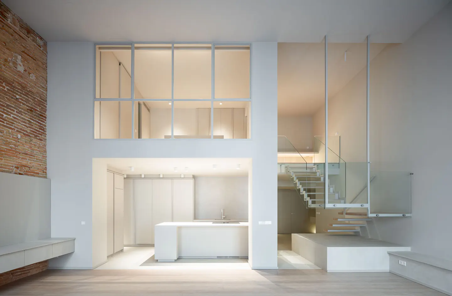 Minimalist loft interior with white walls, wood floors, and a brick accent wall. A modern kitchen and glass-railed staircase are visible.