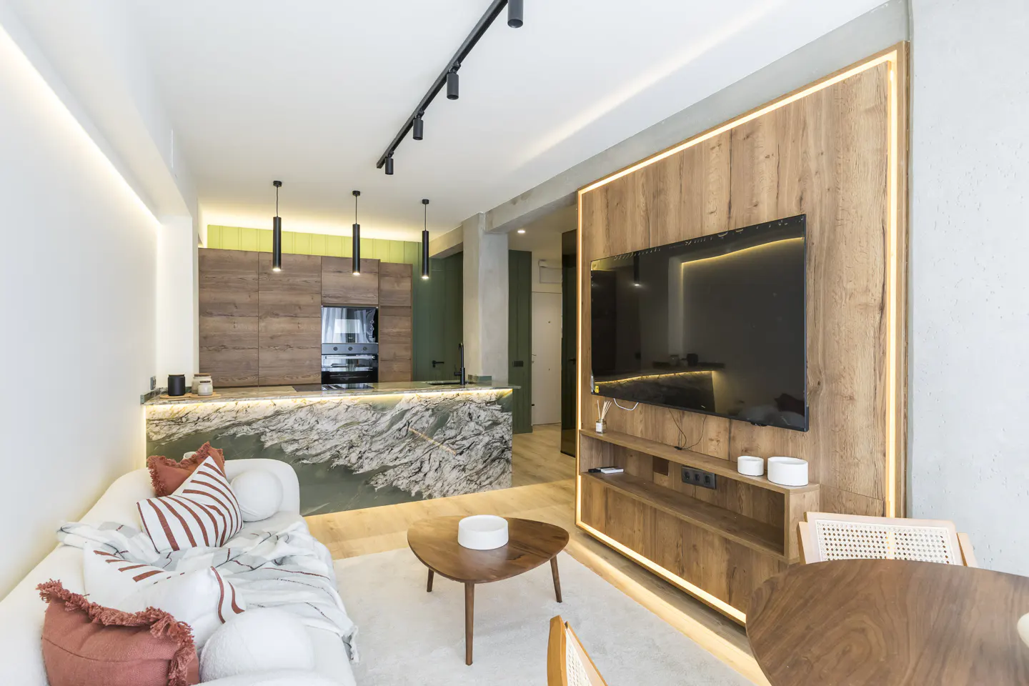Open-concept living space with a white sofa, wood table, and a kitchen island with a marble countertop. A large TV is mounted on a wood-paneled wall.