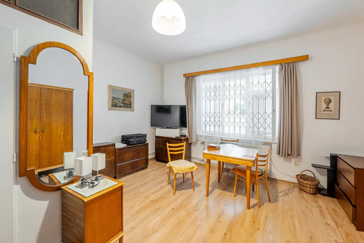 A bright room with wooden furniture, a table with chairs, and a large window with white curtains. A mirror reflects a wardrobe.