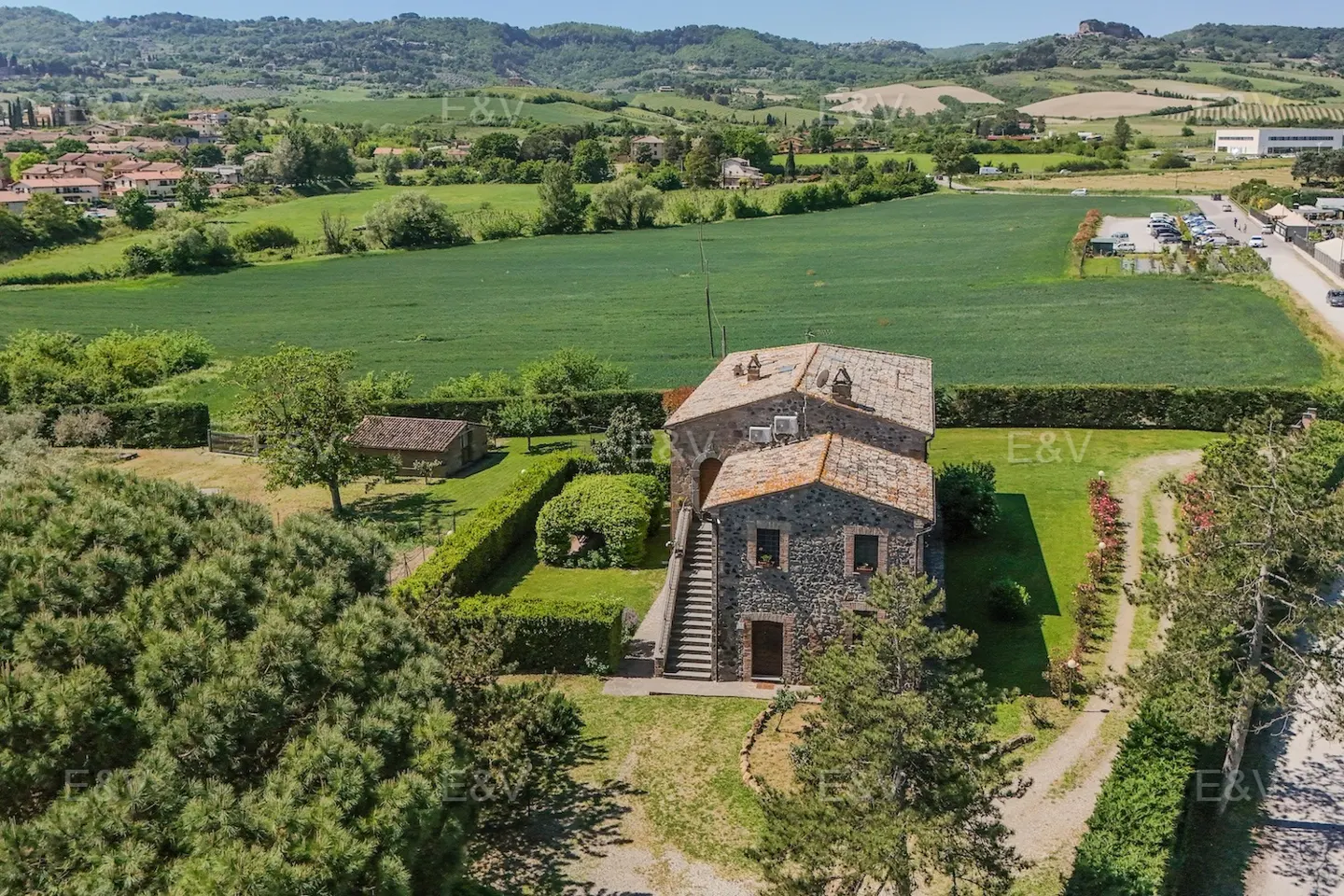 Aerial view of a stone house with a tile roof, surrounded by green lawns, trees, and a green field in the background.