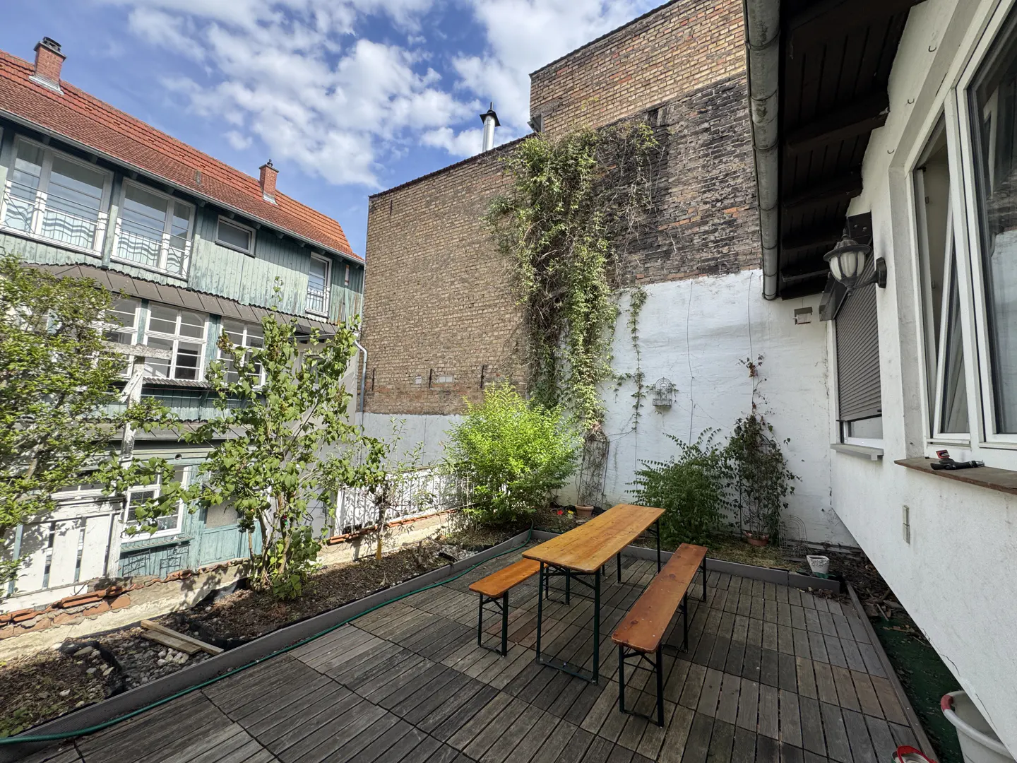 A wooden patio with a picnic table and benches, set against a white wall and a tall, ivy-covered brick building.