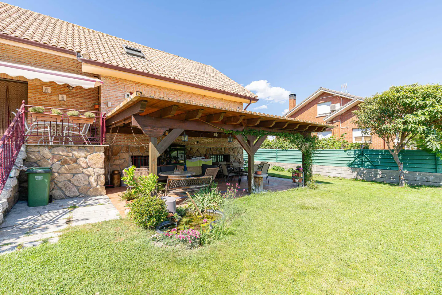 A brick house with a wooden pergola patio, stone base, small pond, and green lawn under a clear blue sky.