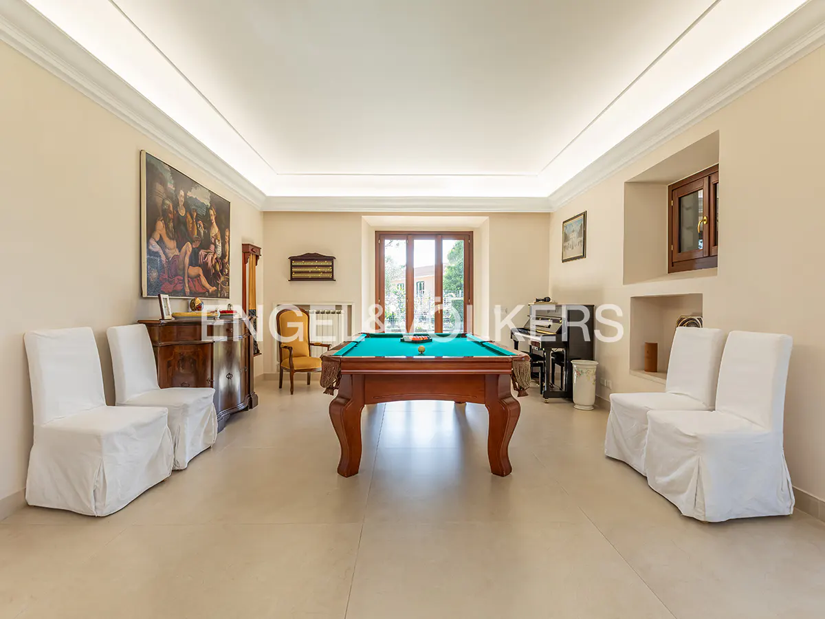 A bright, spacious game room with a pool table, piano, and seating. The walls are light beige, and the floor is tiled.