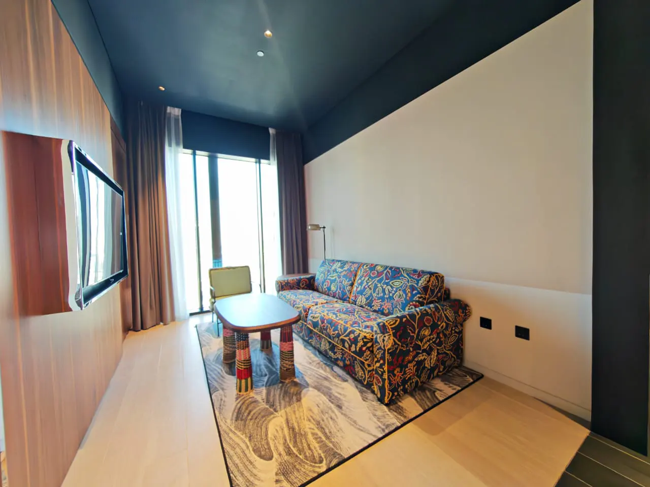 Bright living room with a floral sofa, patterned rug, and a table with colorful legs. A large window with curtains lets in natural light. A TV is mounted on the wall.