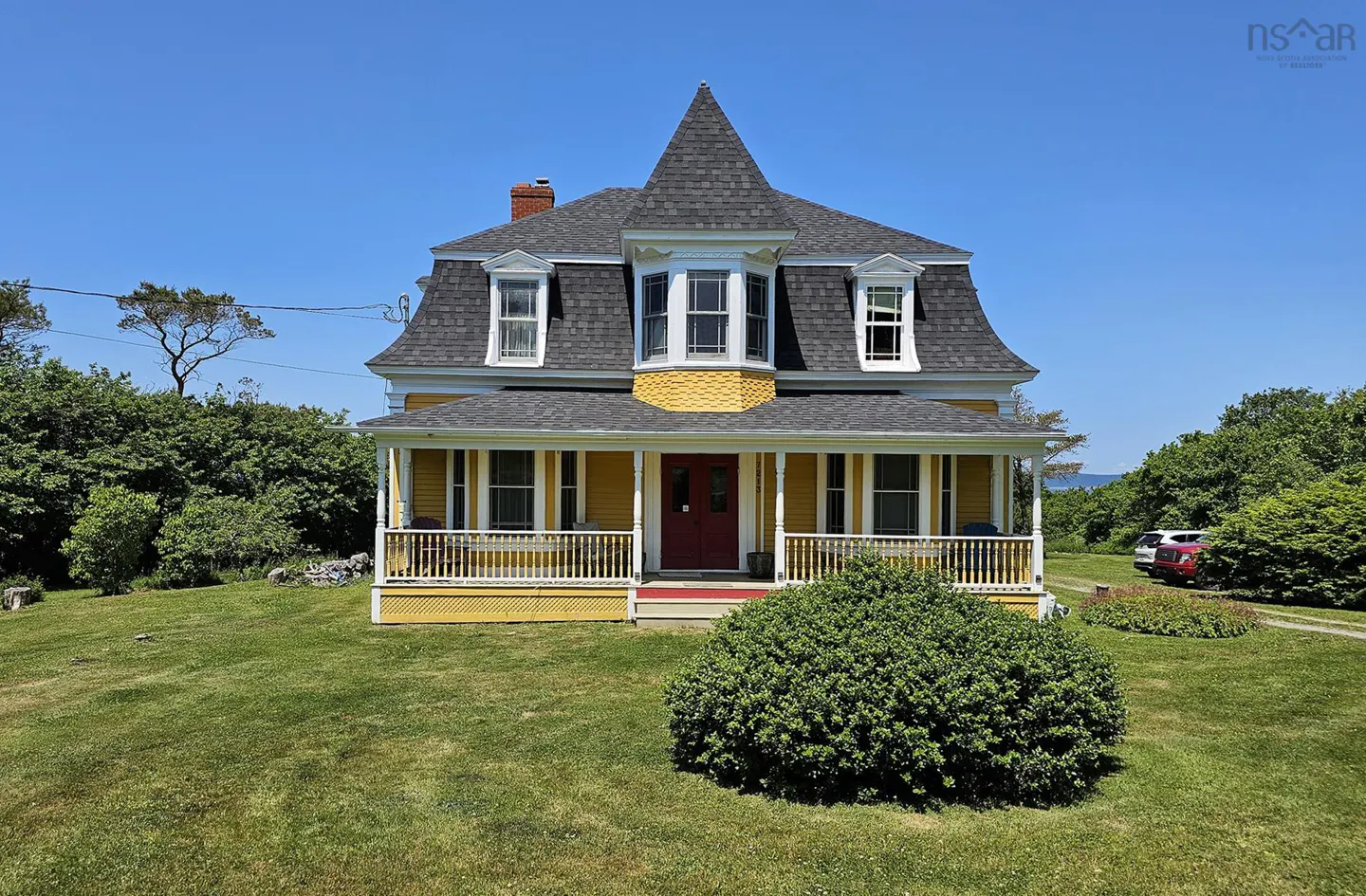 Yellow two-story Victorian house with a red front door, wrap-around porch, and dark shingled roof on a grassy lot.