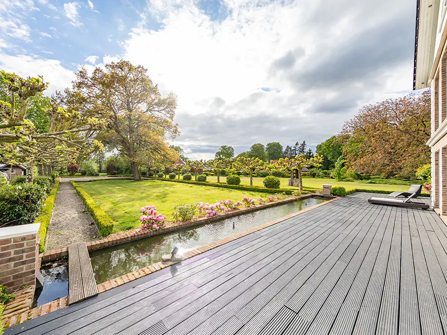 A backyard view from a gray wood deck, showing a long pond with pink flowers, a green lawn, and trees under a cloudy sky.