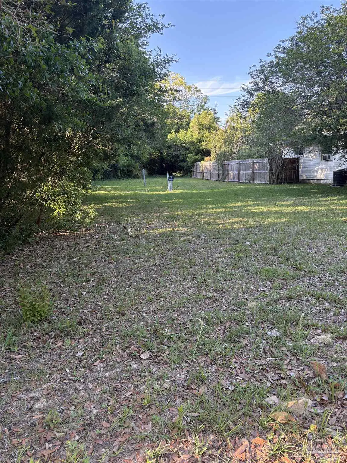 A vacant lot with grass and leaves, bordered by trees and a wooden fence. A white house is visible behind the fence.