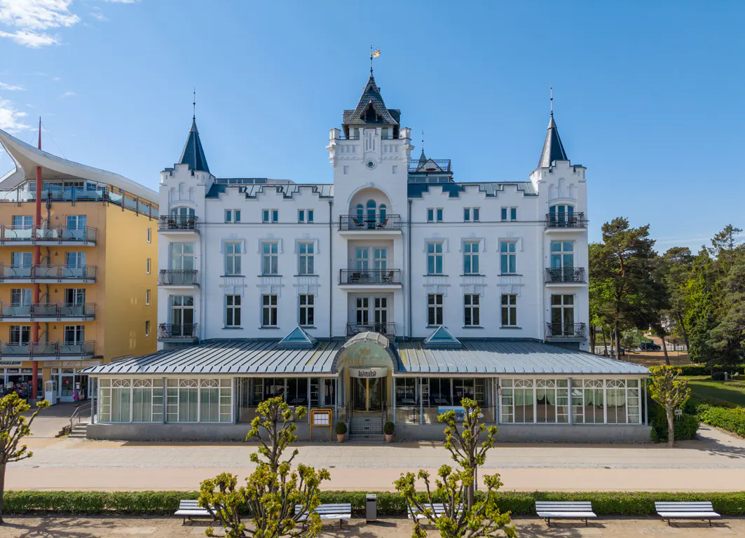 Exterior view of a white, multi-story hotel with turrets and a glass-enclosed restaurant area. A park with benches is in the foreground.