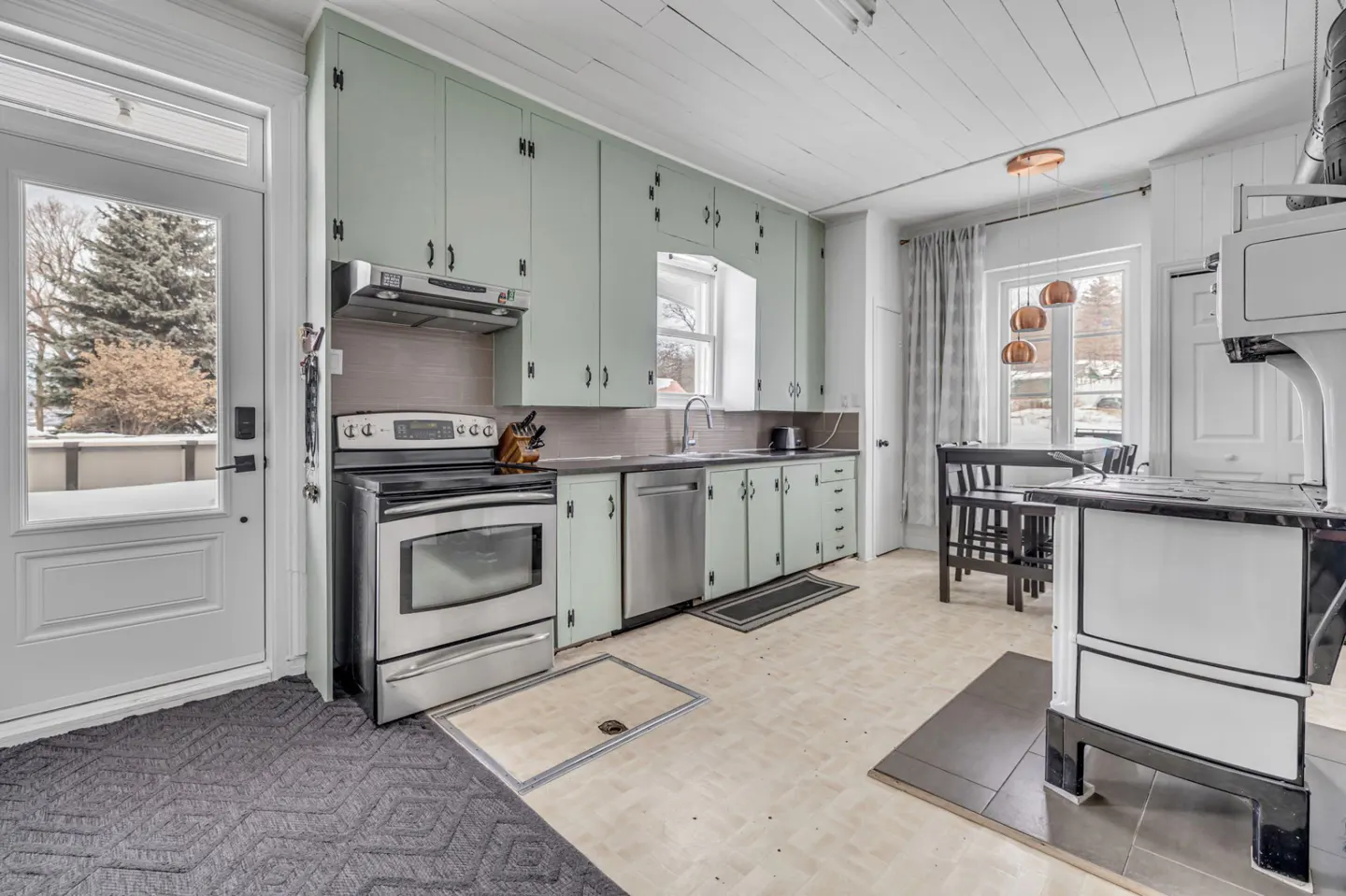 A bright kitchen with light green cabinets, stainless steel appliances, and a white door to the outside. A vintage stove sits near a window with copper pendant lights.