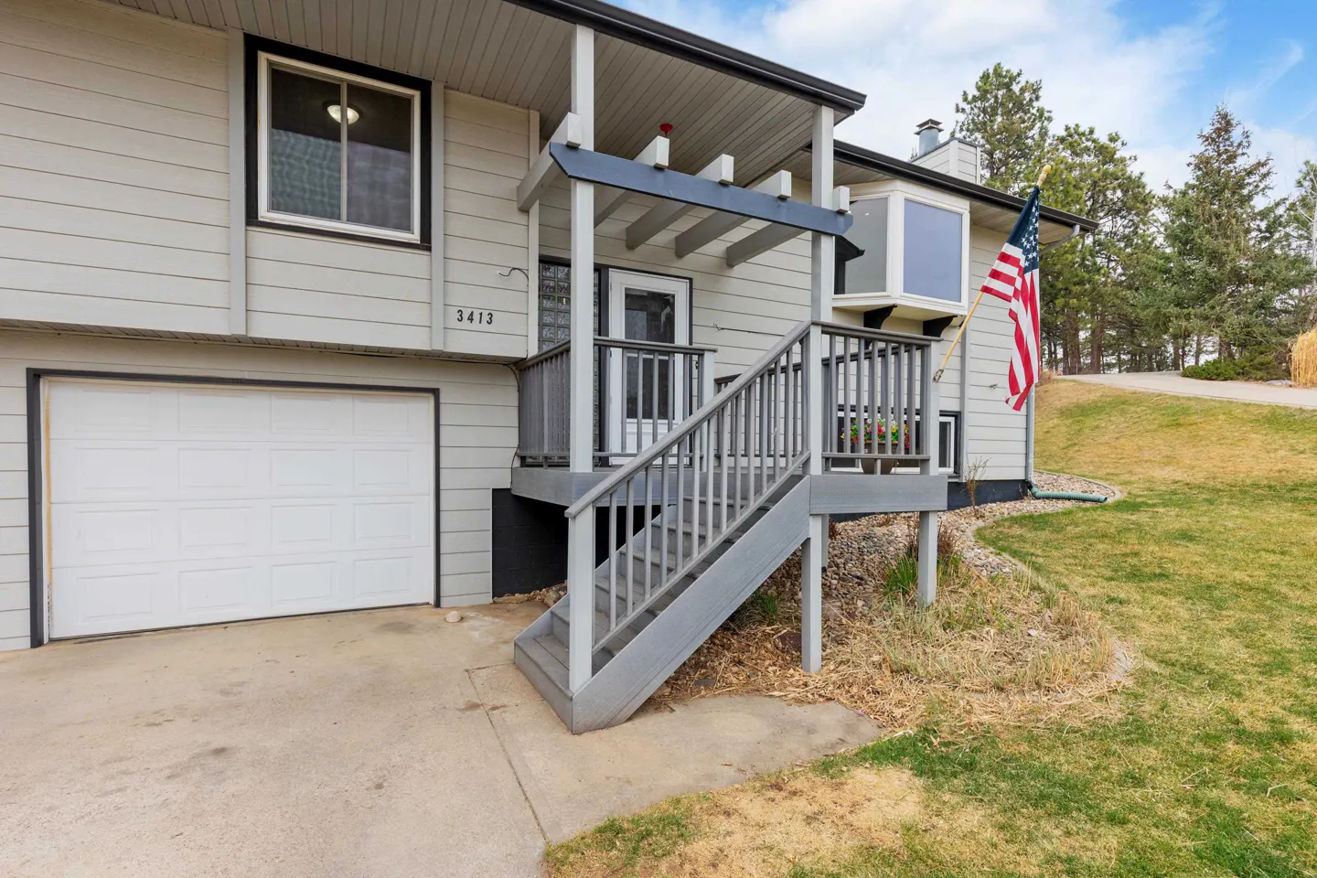 Exterior view of a two-story house with gray siding, a white garage door, and a gray staircase leading to the front door. An American flag is visible.