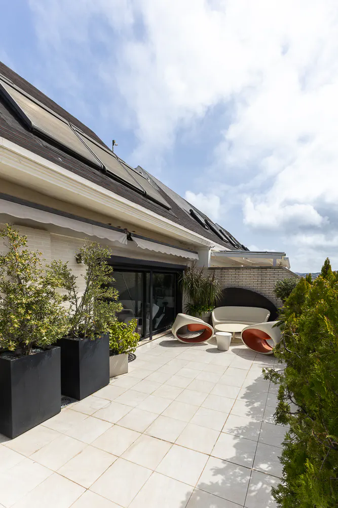 A rooftop patio with white tile, modern white and orange furniture, and potted plants under a cloudy sky.