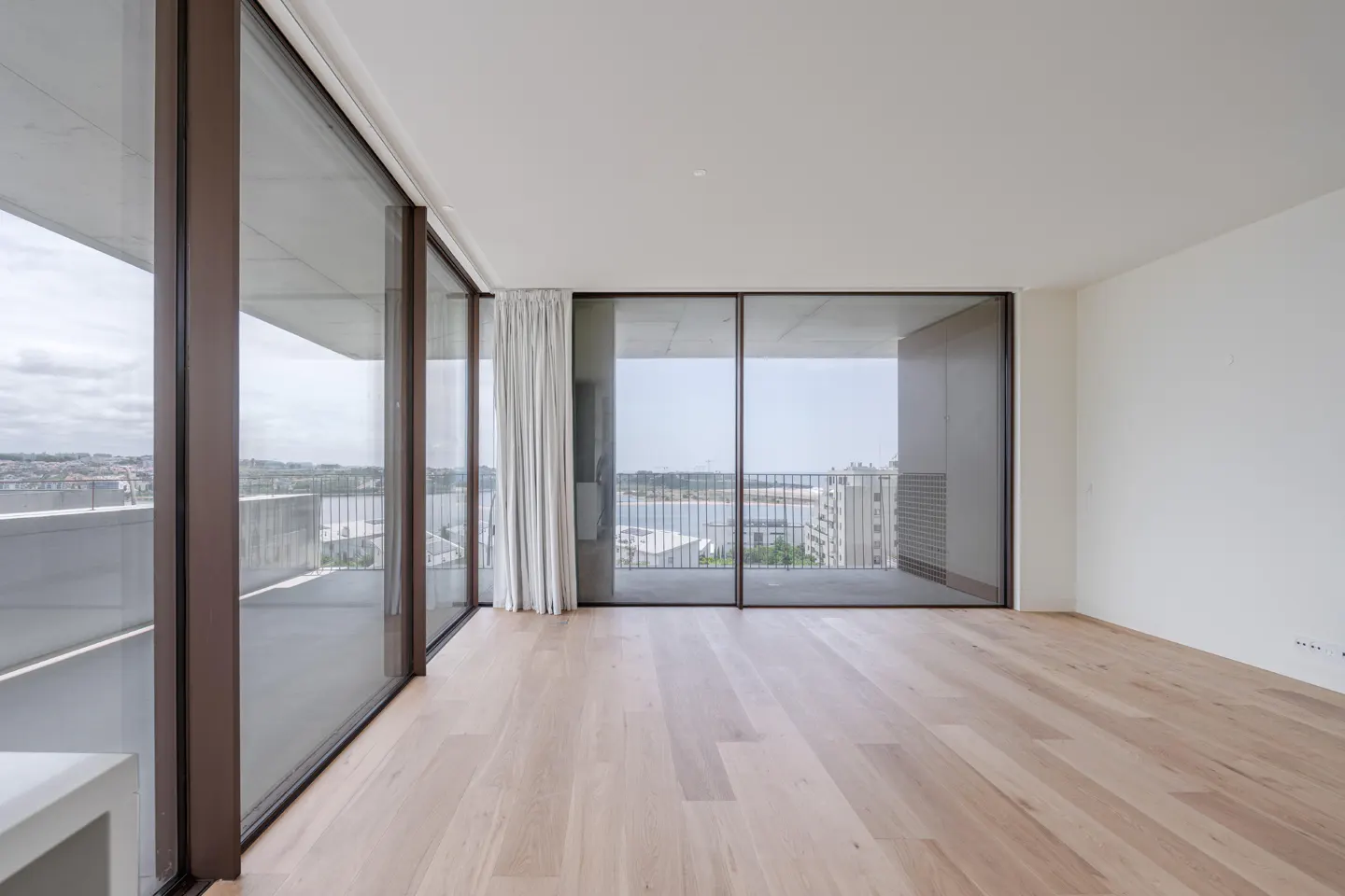 Bright, empty room with light wood floors and white walls. Large sliding glass doors lead to a balcony with a city view.