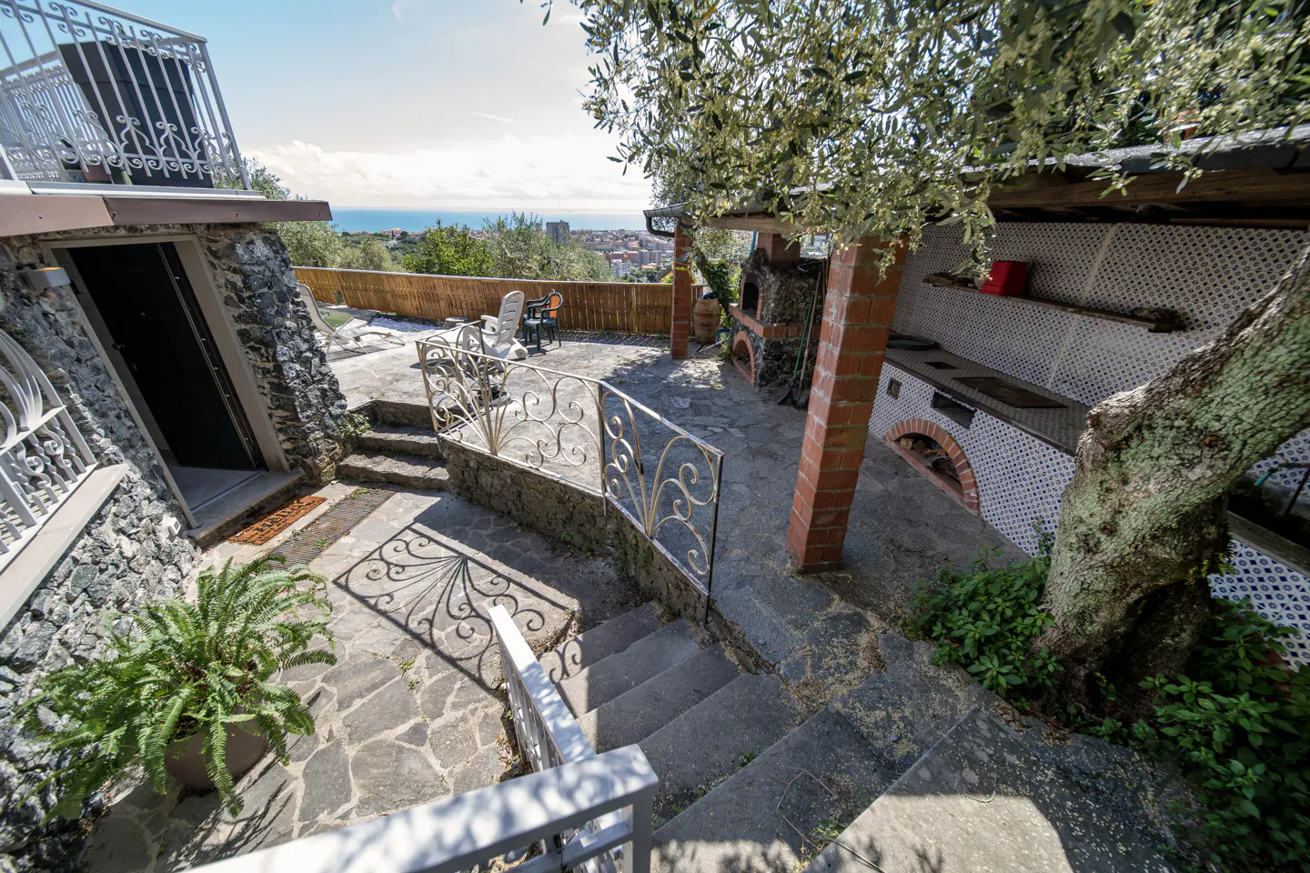 Outdoor patio with stone steps, wrought iron railings, and a built-in outdoor kitchen with brick columns. Distant view of the sea.